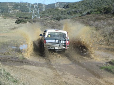 1990 Ford Bronco - pickup truck throws up huge fans of brown mud and water as it powers through a large puddle beneath transmission towers.
