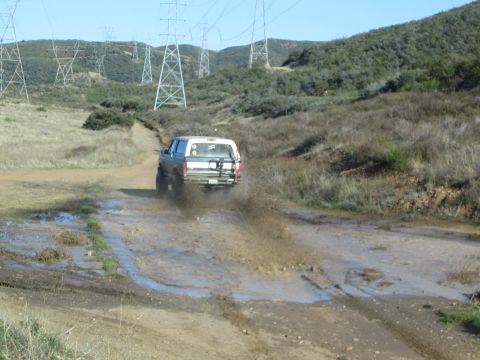 1990 Ford Bronco - pickup truck drives away through a muddy puddle on a dirt trail in a grassy valley lined with power transmission towers.
