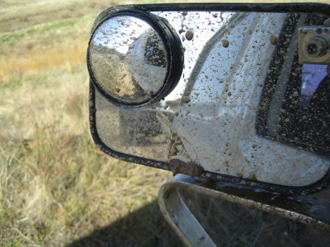 1990 Ford Bronco - Close-up of a white vehicle's side mirror caked with mud spatter, fitted with a round convex blind-spot mirror.