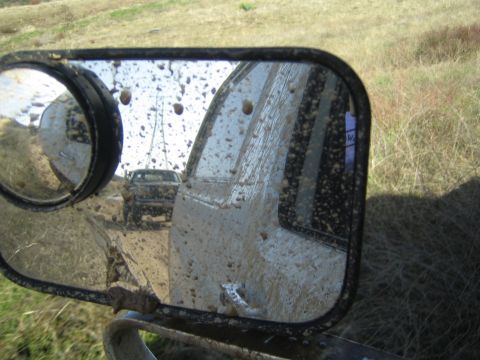 1990 Ford Bronco - mud-spattered side mirror with a round convex insert reflects a distant truck and grassy field behind the vehicle.