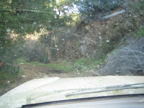 1990 Ford Bronco - Looking through a speckled windshield over a white hood at a rocky roadcut and trees along a wooded dirt trail.