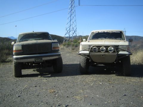 1990 Ford Bronco - Two lifted off-road trucks, one with a billet grille and one with a tube brush guard and round lights, parked side by side beneath a transm…