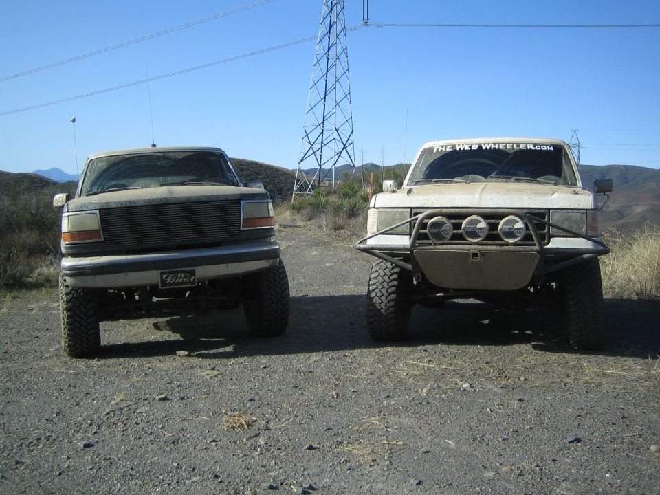 Front view of two lifted off-road trucks parked side by side under transmission tower