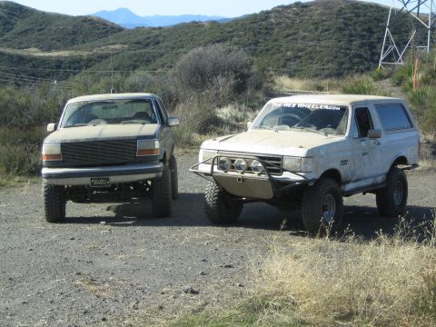 1990 Ford Bronco - Two lifted Ford full-size SUVs, one tan and one white with a tube bumper, parked on a dirt road among green chaparral hills.