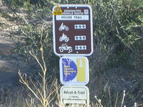 1990 Ford Bronco - Brown Designated Vehicle Route sign for House Trail above a Back Country Discovery Trails marker and Adopt-A-Trail plaque in dry brush.