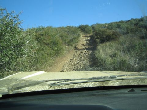 1990 Ford Bronco - View through a dusty windshield over a vehicle hood up a steep, rocky off-road trail climbing a brush-covered hill.