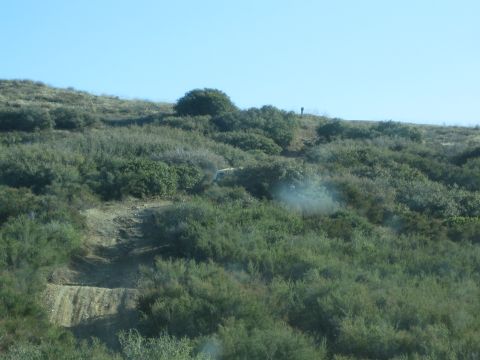 1990 Ford Bronco - light-colored vehicle sits on a switchback trail across a green brush-covered hillside, kicking up faint dust.