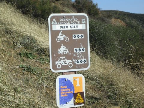 1990 Ford Bronco - Brown Designated Vehicle Route sign for Deer Trail with difficulty icons, above a Back Country Discovery Trails marker on a dry grassy slop…
