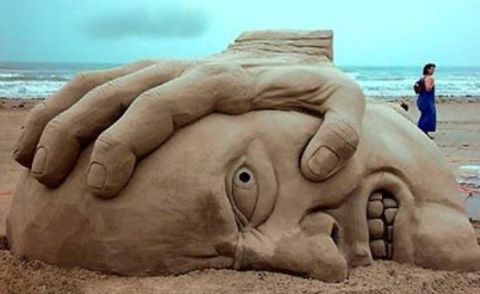 1989 Ford Bronco - A large sand sculpture depicts a hand pressing down on a head, with the ocean and a distant figure on the beach.