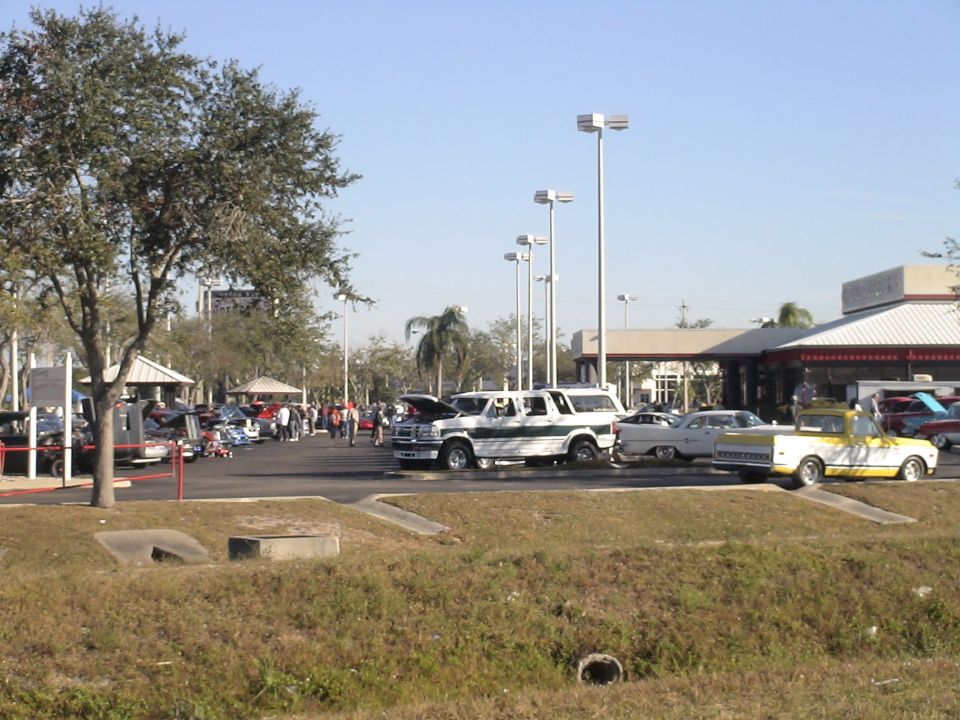 Car show lot with a white-and-green full-size Ford SUV conversion, hood open, among classic cars