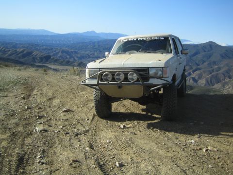 1990 Ford Bronco - dirty white lifted off-road SUV with a tube bumper and three driving lights sits on a dirt ridge with mountain ranges behind.
