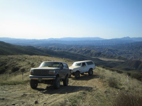 1990 Ford Bronco - tan pickup-style Ford and a white SUV, both lifted, parked on a dirt ridge above a sweeping view of distant hills.