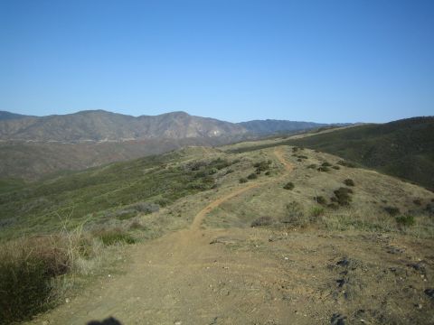 1990 Ford Bronco - dirt trail winds along a brushy ridgeline toward distant blue mountains under a clear sky.