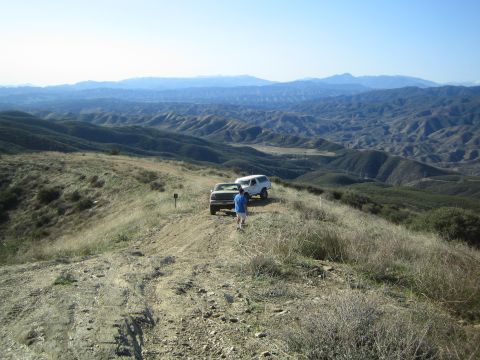 1990 Ford Bronco - person in a blue shirt walks toward a white SUV parked on a dirt ridge overlooking a vast expanse of rolling hills.