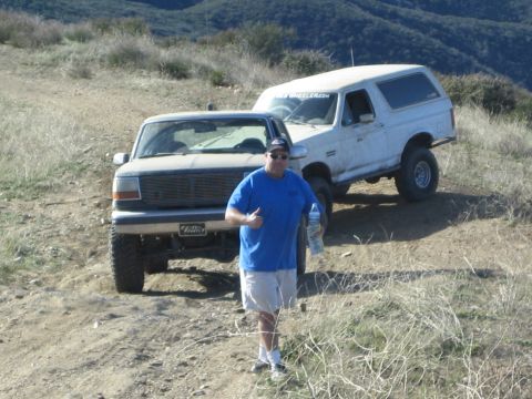 1990 Ford Bronco - man in a blue shirt gives a thumbs-up holding a water bottle beside a tan and a white lifted Ford SUV on a hillside trail.