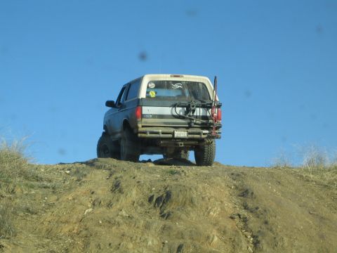 1990 Ford Bronco - Rear view of a lifted Ford SUV with a rear tire carrier cresting a dirt hill against a clear blue sky.