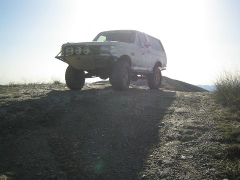 1990 Ford Bronco - white lifted Ford SUV with a tube bumper and driving lights stands backlit on a dirt ridge at low sun.