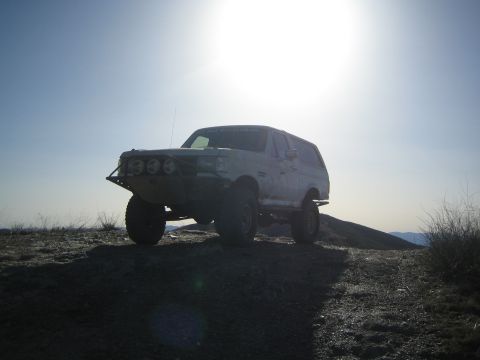 1990 Ford Bronco - white lifted Ford SUV with a tube bumper sits on a hilltop, strongly backlit by the sun with lens flare.