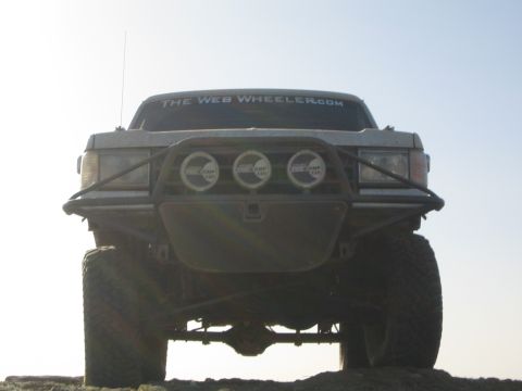 1990 Ford Bronco - Shot from below against a bright sky, a lifted vehicle shows its front tube bumper, three round driving lights, and a tall whip antenna.