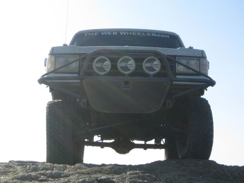 1990 Ford Bronco - lifted off-road vehicle perched on a rock outcrop is shown from a low front angle, with three round lights mounted in its tube bumper.