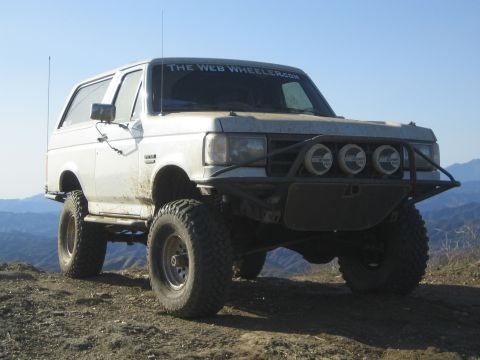 1990 Ford Bronco - white lifted two-door SUV with a windshield banner and front tube bumper sits on a dirt ridge with mountains in the background.