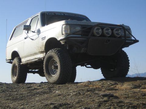 1990 Ford Bronco - muddy white lifted SUV with large tires and a front tube bumper holding three driving lights rests on a dirt hilltop under a clear sky.