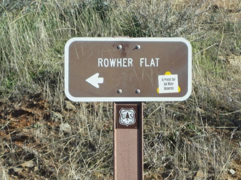 1990 Ford Bronco - brown US Forest Service directional sign reading Rowher Flat with a left arrow stands on a post against a dry, rocky hillside.