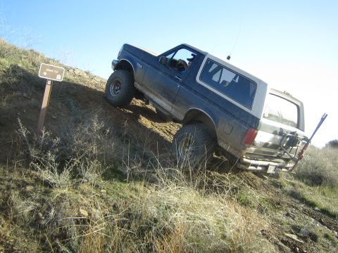 1990 Ford Bronco - dark lifted two-door SUV climbs a steep grassy embankment beside a trail marker post under a clear blue sky.