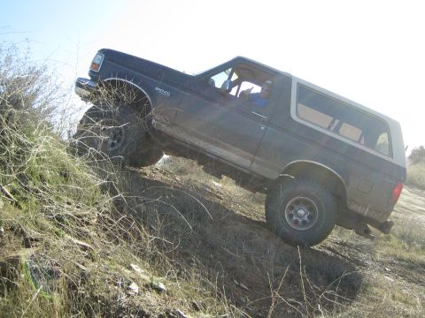1990 Ford Bronco - dark lifted two-door SUV with a visible driver climbs a brushy dirt slope, body tilted on the off-camber terrain.