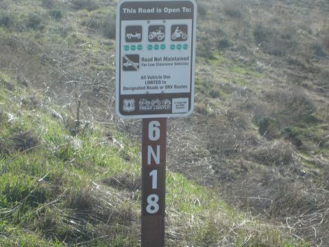 1990 Ford Bronco - brown Forest Service sign listing permitted off-road vehicles is mounted on a post marked 6N18 against a dry grassy hillside.