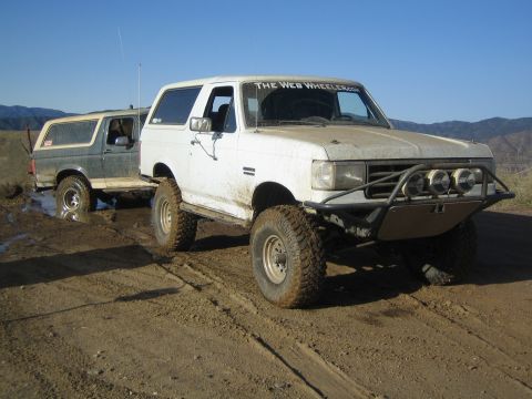 1990 Ford Bronco - Two lifted two-door SUVs, one white with a tube bumper and one darker, sit side by side on a muddy trail with mountains beyond.