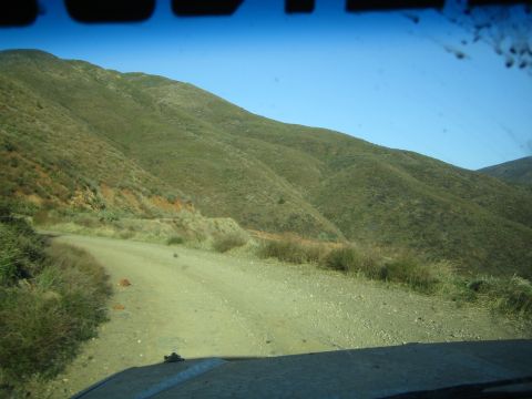 1990 Ford Bronco - Seen through a dusty windshield, a dirt road curves along the base of grassy green hills under a clear blue sky.