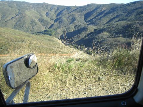 1990 Ford Bronco - Looking past a vehicle's worn side mirror through the window, green hills and a steep canyon slope fill the view.