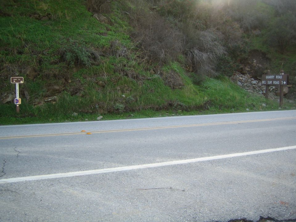 Two-lane paved road below a green hillside with small brown directional signs