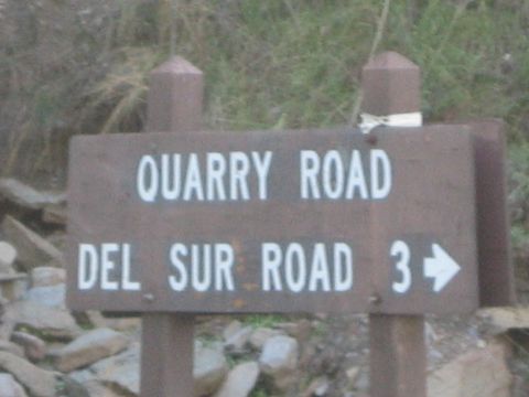 1990 Ford Bronco - brown wooden sign reads Quarry Road above Del Sur Road 3 with a right arrow, mounted on two posts amid rocks and brush.