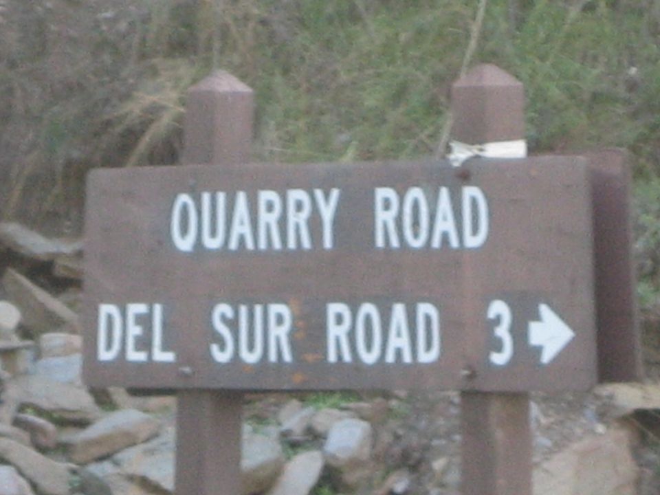 Brown wooden sign reading Quarry Road and Del Sur Road 3 with a right arrow