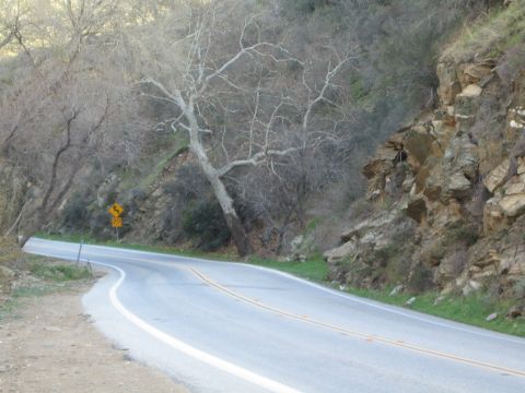 1990 Ford Bronco - two-lane mountain road curves past rocky cliffs and leafless trees, with a yellow warning sign on the left shoulder.