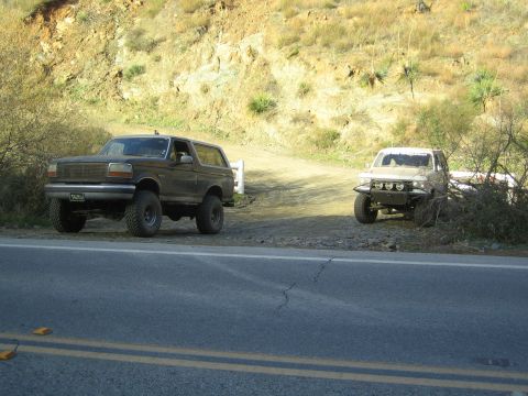 1990 Ford Bronco - Two lifted SUVs are parked on the shoulder of a paved two-lane road at the foot of a dusty dirt hillside.