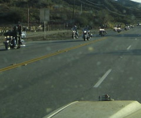 1990 Ford Bronco - line of motorcycles rides down a rural highway beside dry hills, seen through a spotted, dirty windshield.