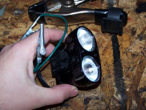1986 Ford Bronco - A hand holds a black dual-bulb underhood light assembly connected to jumper cables on a wooden surface.
