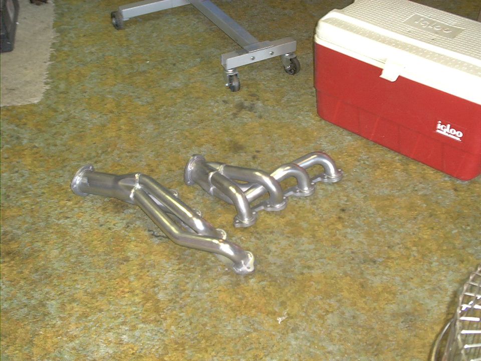 Two chrome exhaust headers on a carpeted floor next to a red Igloo cooler and a metal stand