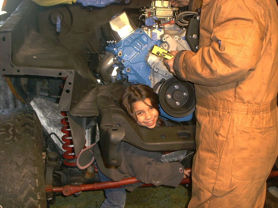Child peeking out from under engine during vehicle repair. Adult in tan jumpsuit works on engine