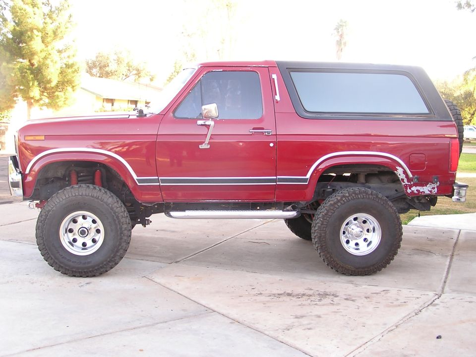 Lifted red 1986 Ford Bronco with black top and large off-road tires, side view on a driveway