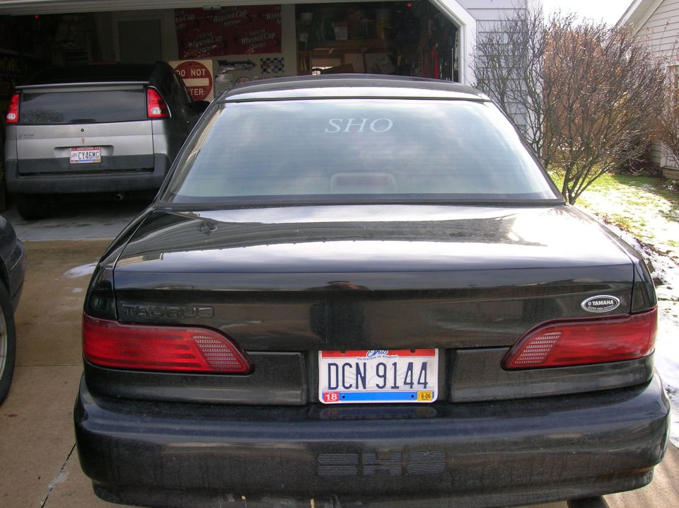 Rear view of a black 1992 Ford Taurus SHO with a license plate DCN 9144. Another vehicle is parked in the garage behind it