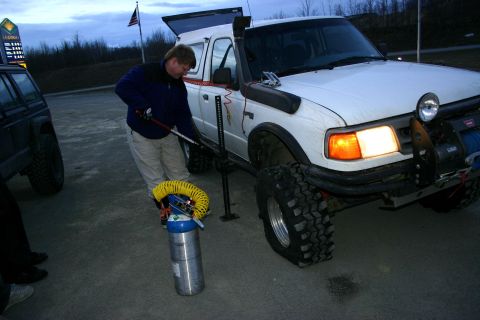 2003 Jeep TJ - A man uses a jack to lift a white Ford Ranger pickup truck while another vehicle is parked nearby.