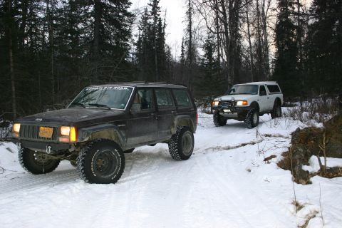 2003 Jeep TJ - A dark 2003 Jeep Cherokee XJ with a "KODIAK OFF-ROAD" sticker drives through the snow, followed by a white pickup truck.