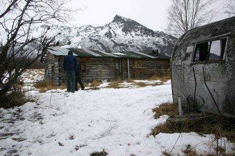 2003 Jeep TJ - An off-road vehicle with its headlights illuminated is visible in a snowy landscape near a rustic log cabin and an old trailer.