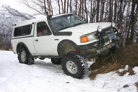 2003 Jeep TJ - A white pickup truck navigates a snowy, wooded area with its tires on a dirt mound.
