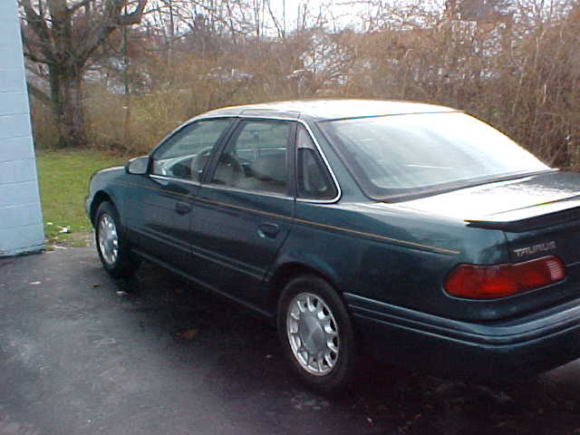 Dark green Ford Taurus sedan, rear driver-side view, parked on driveway near blue building