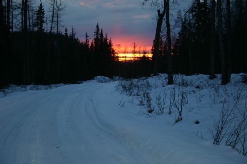 2003 Jeep TJ - A winter road winds through a dark forest as the sun sets, casting a colorful glow on the horizon.